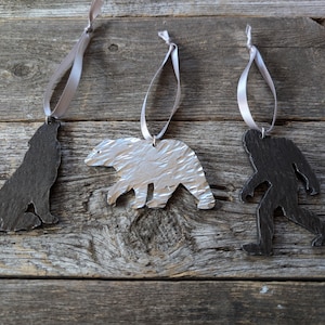 May include: Three wooden ornaments in the shape of a wolf, a bear, and a Bigfoot. The ornaments are hanging from silver ribbon.