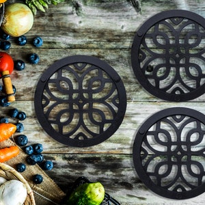 Puede incluir: Tres salvamanteles de metal negro con un diseño decorativo intrincado. Los salvamanteles son redondos y tienen un borde elevado. Están dispuestos en fila sobre una superficie de madera.