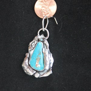 May include: A silver pendant with a turquoise stone set in a textured silver design. The pendant is attached to a silver bail.