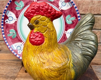 Peut inclure: Une figurine de coq en céramique avec un corps jaune, une crête et des barbillons rouges et une queue grise. Le coq est debout sur une surface en bois devant une assiette rouge et verte avec un motif de coq blanc.
