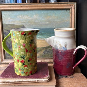 May include: Two ceramic pitchers, one green with a floral pattern and one with a white and burgundy glaze. The pitchers are sitting on a stack of old books in front of a framed painting of a seascape.