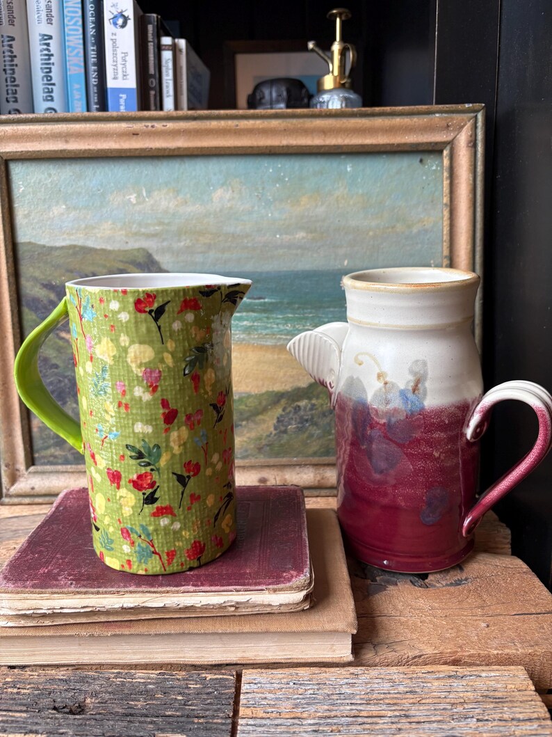 May include: Two ceramic pitchers, one green with a floral pattern and one with a white and burgundy glaze. The pitchers are sitting on a stack of old books in front of a framed painting of a seascape.