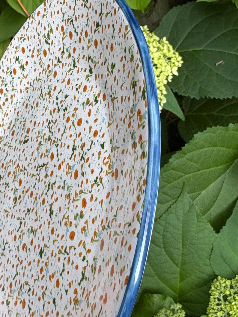 Może przedstawiać: Biały ceramiczny talerz z niebieską obw&oacute;dką. Powierzchnia talerza pokryta jest wzorem małych pomarańczowych, zielonych i niebieskich kształt&oacute;w przypominających kwiaty. Talerz jest częściowo widoczny.