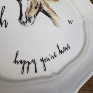 May include: White ceramic plate with a scalloped edge, featuring a sketched horse head in brown and tan. The plate has the handwritten text "happy you're here" in black cursive script. A decorative piece for the home.