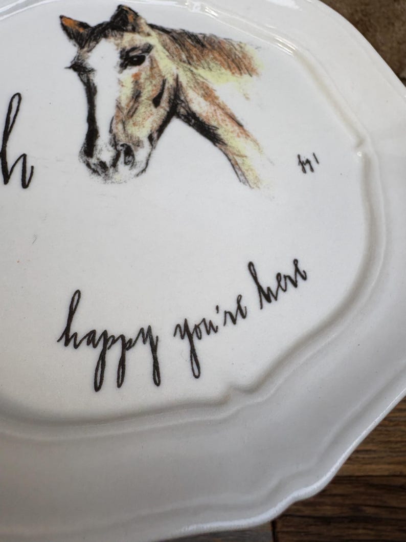 May include: White ceramic plate with a scalloped edge, featuring a sketched horse head in brown and tan. The plate has the handwritten text "happy you're here" in black cursive script. A decorative piece for the home.