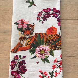 Puede incluir: Pa&ntilde;o de cocina blanco con un dise&ntilde;o de tigre con rayas naranjas y negras, rodeado de flores rosas y moradas y un colibr&iacute;. El pa&ntilde;o tiene un tema floral y animal.