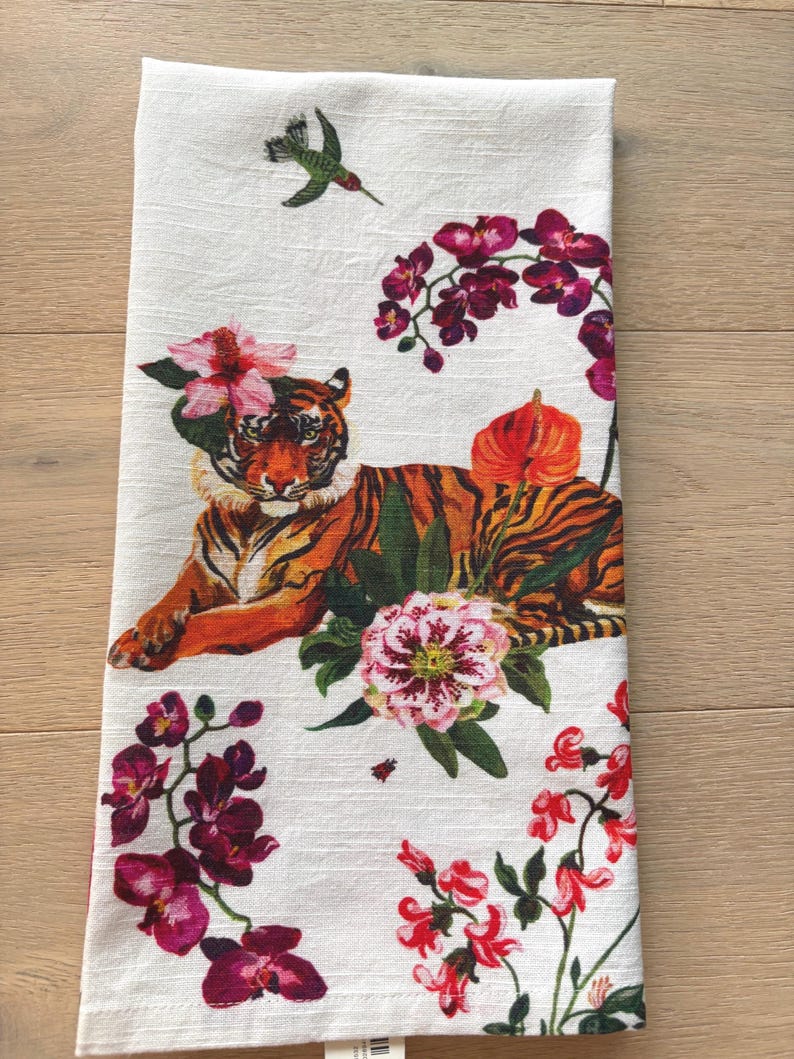 Puede incluir: Pa&ntilde;o de cocina blanco con un dise&ntilde;o de tigre con rayas naranjas y negras, rodeado de flores rosas y moradas y un colibr&iacute;. El pa&ntilde;o tiene un tema floral y animal.