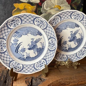May include: Two blue and white decorative plates with a bird and landscape design. The plates have a scalloped edge and are displayed on brass stands. The plates are on a rustic wooden surface, with a floral painting in the background.