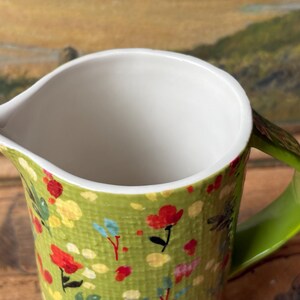 May include: A green ceramic pitcher with a white interior and a green handle. The pitcher is decorated with a floral pattern in shades of red, yellow, and blue.