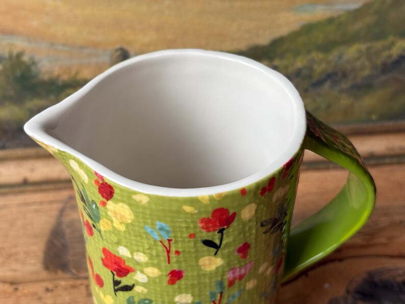 May include: A green ceramic pitcher with a white interior and a green handle. The pitcher is decorated with a floral pattern in shades of red, yellow, and blue.