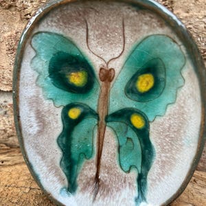 Puede incluir: Un plato de cerámica con un diseño de mariposa pintado. La mariposa es verde con detalles amarillos y marcas marrones. El plato es de forma ovalada y tiene un fondo blanco.