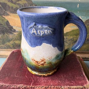 Puede incluir: Una taza de cerámica hecha a mano con un exterior azul, con un diseño de paisaje blanco y verde. La taza tiene un asa y la palabra "Aspen" está grabada en relieve en la parte delantera. La taza está sobre un libro rojo.