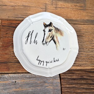 May include: White ceramic decorative plate with a scalloped edge. The plate features a hand-drawn horse head illustration in brown and tan, with the words "Hh" and "happy you're here" in black script.
