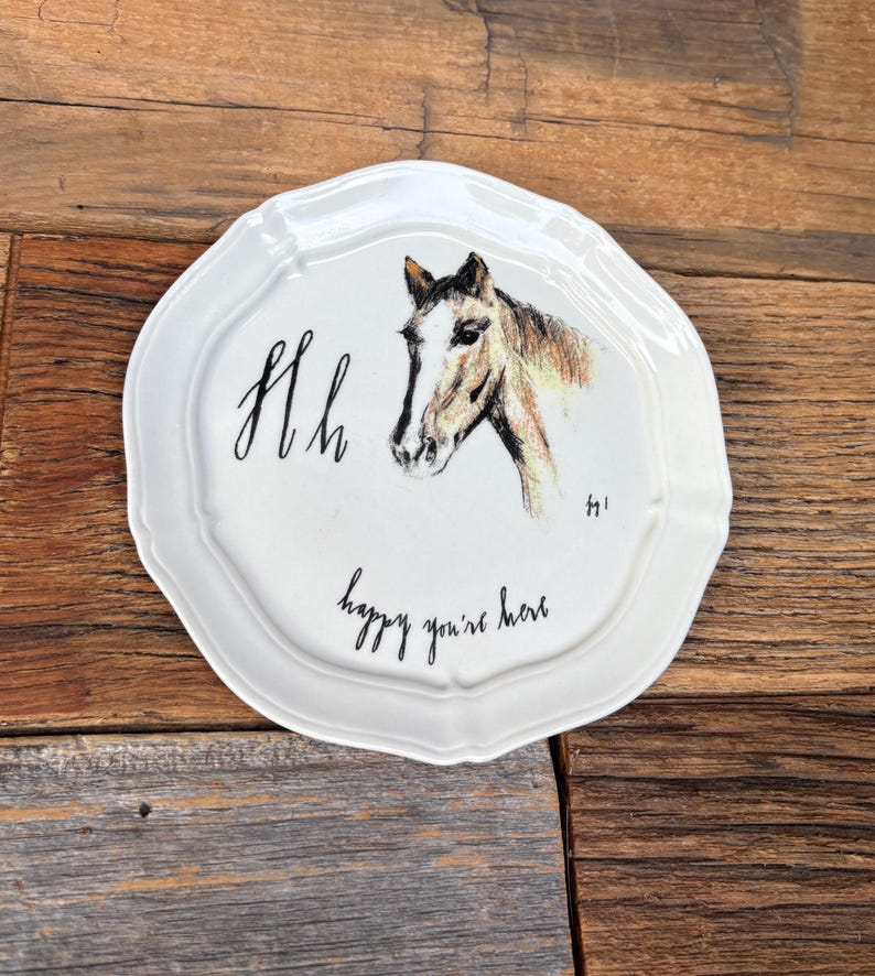 May include: White ceramic decorative plate with a scalloped edge. The plate features a hand-drawn horse head illustration in brown and tan, with the words "Hh" and "happy you're here" in black script.