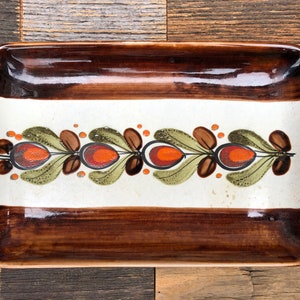 Puede incluir: Un plato rectangular de cerámica con un borde marrón y un centro blanco. El plato presenta un diseño decorativo de hojas verdes, puntos marrones y acentos naranja y rojos.