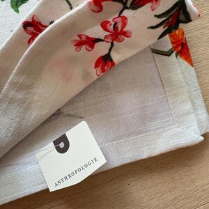 Puede incluir: Tela de lino blanquecina con un estampado floral de flores rojas, rosas y naranjas y hojas verdes. La tela est&aacute; doblada, revelando una etiqueta con la palabra "ANTHROPOLOGIE".