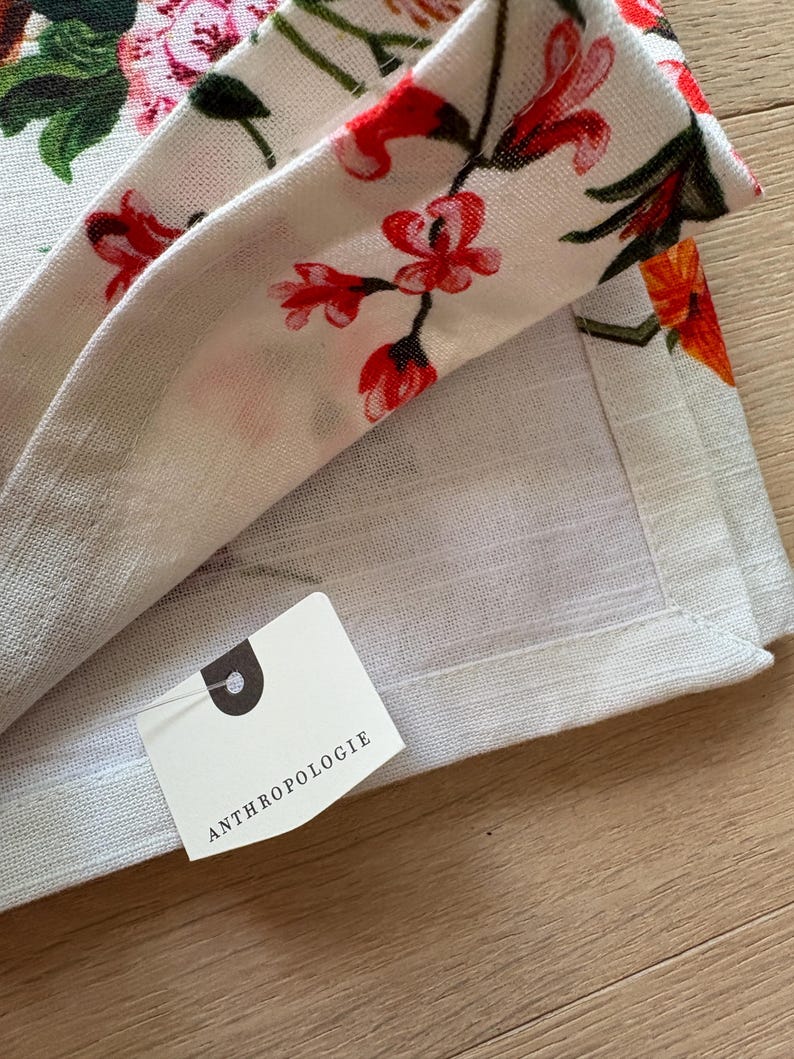 Puede incluir: Tela de lino blanquecina con un estampado floral de flores rojas, rosas y naranjas y hojas verdes. La tela est&aacute; doblada, revelando una etiqueta con la palabra "ANTHROPOLOGIE".