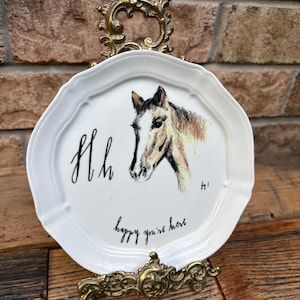 May include: White decorative plate with a horse head illustration, the letters "Hh", and the phrase "happy you're here". The plate is displayed on a decorative gold-colored stand.