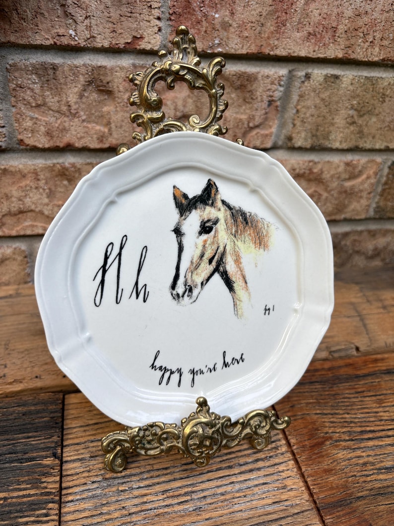 May include: White decorative plate with a horse head illustration, the letters "Hh", and the phrase "happy you're here". The plate is displayed on a decorative gold-colored stand.