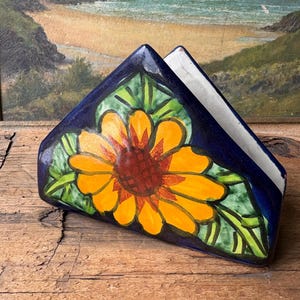 May include: A ceramic napkin holder featuring a sunflower design. The holder is triangular, with a dark blue exterior and a white interior. The sunflower has yellow petals and a brown centre, surrounded by green leaves.