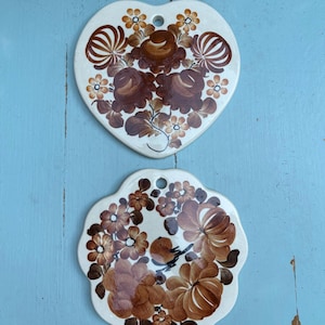 Puede incluir: Dos piezas decorativas de cerámica. Una tiene forma de corazón, la otra de flor. Ambas son blancas con diseños florales marrones. La pieza en forma de corazón tiene un agujero en la parte superior. La pieza en forma de flor tiene un diseño de pájaro.