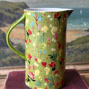 May include: A green ceramic pitcher with a floral pattern. The pitcher has a white rim and a green handle.