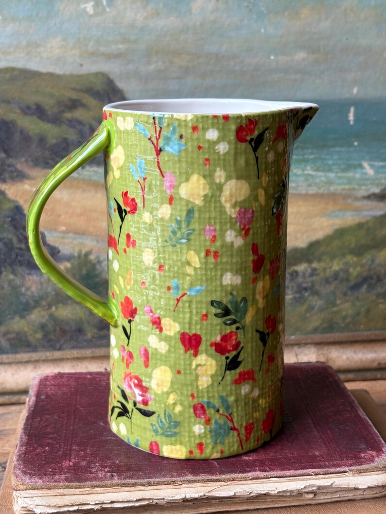 May include: A green ceramic pitcher with a floral pattern. The pitcher has a white rim and a green handle.