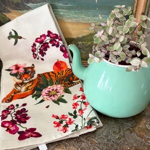 Puede incluir: Un pa&ntilde;o de cocina decorativo con un dise&ntilde;o de tigre y flores, junto a una tetera verde menta con una planta en su interior. La toalla presenta un tigre con flores y un colibr&iacute;. La tetera est&aacute; sobre una bandeja plateada.