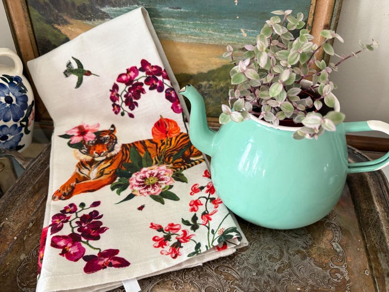Puede incluir: Un pa&ntilde;o de cocina decorativo con un dise&ntilde;o de tigre y flores, junto a una tetera verde menta con una planta en su interior. La toalla presenta un tigre con flores y un colibr&iacute;. La tetera est&aacute; sobre una bandeja plateada.