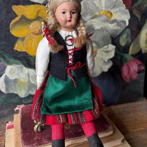 May include: A vintage doll with blonde braids and a gold crown. The doll wears a white blouse, black vest with red lacing, a green apron, and a red and plaid skirt. Red stockings and black shoes complete the outfit.