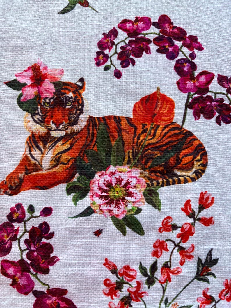 Puede incluir: Textil blanco con una ilustraci&oacute;n detallada de un tigre adornado con flores. El tigre tiene rayas naranjas y negras, con flores rosas y rojas y hojas verdes. Tambi&eacute;n se ve un colibr&iacute;.