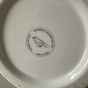 以下が含まれることがあります： 白いセラミック皿。円形のエンブレムには鳥のシルエットと「MICROWAVE & DISHWASHER SAFE」と「MADE IN CHINA」の文字が描かれています。 皿の表面はわずかにざらざらしています。