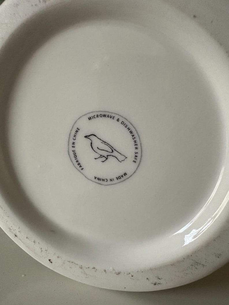 以下が含まれることがあります： 白いセラミック皿。円形のエンブレムには鳥のシルエットと「MICROWAVE & DISHWASHER SAFE」と「MADE IN CHINA」の文字が描かれています。 皿の表面はわずかにざらざらしています。