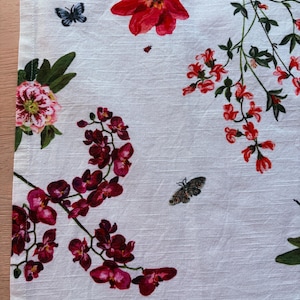 Puede incluir: Pa&ntilde;o de cocina de lino blanco con un dise&ntilde;o floral. El dise&ntilde;o incluye flores rojas, rosas y moradas, hojas verdes y mariposas. Las flores incluyen tulipanes, orqu&iacute;deas y otras flores. El pa&ntilde;o tiene un borde con dobladillo.