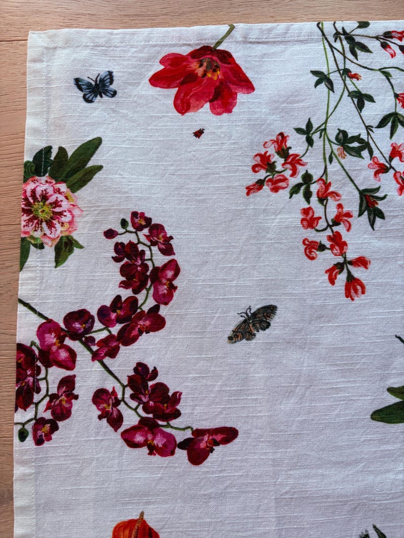 Puede incluir: Pa&ntilde;o de cocina de lino blanco con un dise&ntilde;o floral. El dise&ntilde;o incluye flores rojas, rosas y moradas, hojas verdes y mariposas. Las flores incluyen tulipanes, orqu&iacute;deas y otras flores. El pa&ntilde;o tiene un borde con dobladillo.