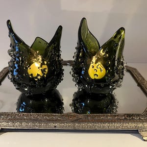 Peut inclure: Deux bougeoirs en verre vert foncé avec une surface texturée et un accent doré, présentés sur un plateau miroir avec un cadre argenté orné. Les bougeoirs ont une forme unique et organique.