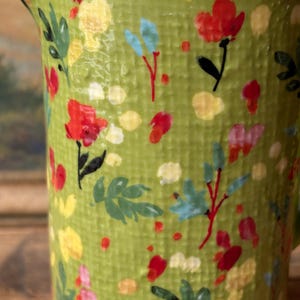May include: A green ceramic pitcher with a floral pattern. The pitcher has a textured surface and features red, yellow, and blue flowers.