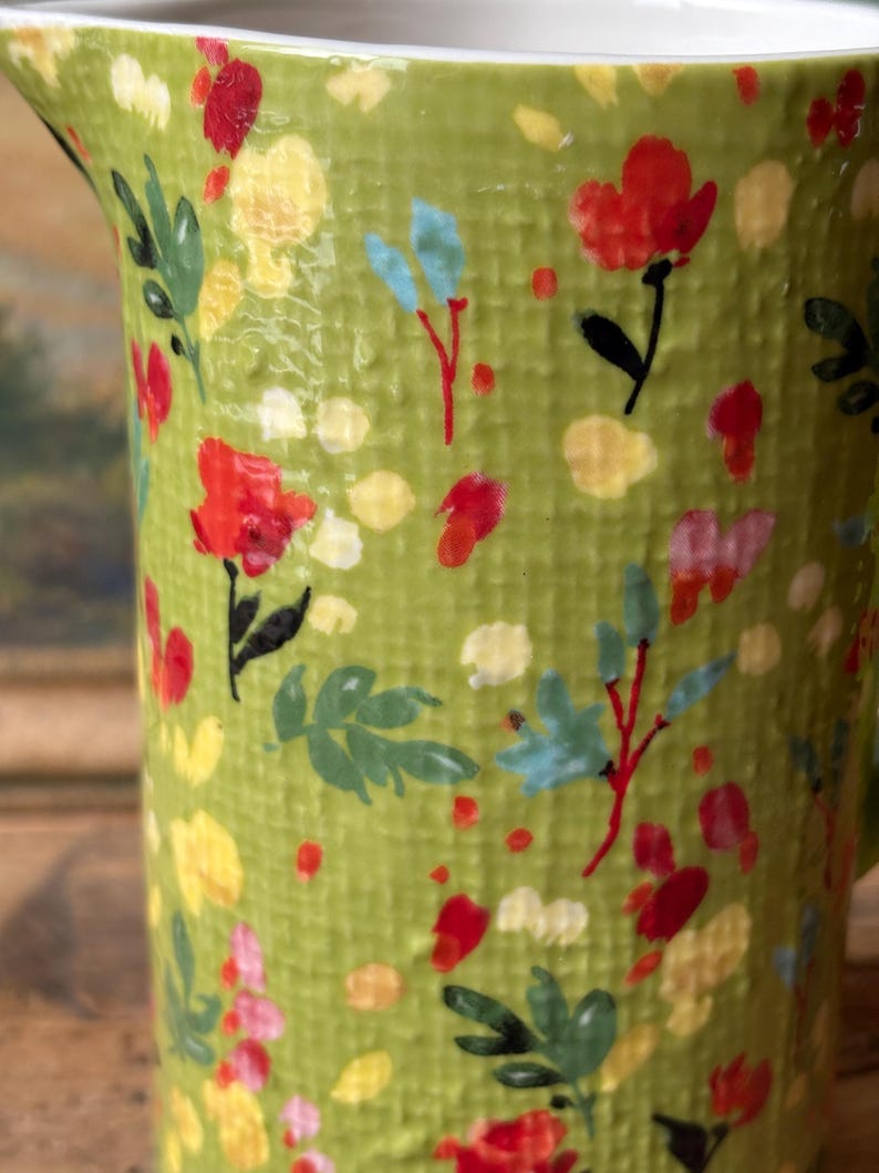 May include: A green ceramic pitcher with a floral pattern. The pitcher has a textured surface and features red, yellow, and blue flowers.
