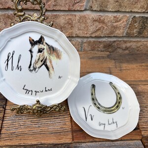 May include: Two white decorative plates with scalloped edges. One plate features a horse head and the text "happy you're here". The other plate has a horseshoe and the text "very lucky".