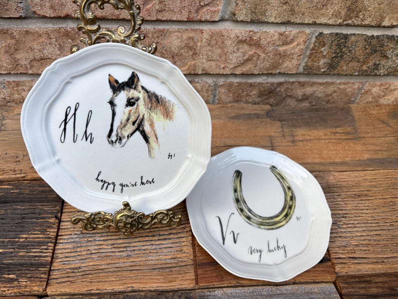 May include: Two white decorative plates with scalloped edges. One plate features a horse head and the text "happy you're here". The other plate has a horseshoe and the text "very lucky".