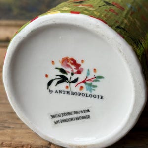 May include: A white ceramic vase with a green and red floral pattern. The vase is marked "by Anthropologie" and "Dishwasher & Microwave Safe. Made in China/Fabriqu&eacute; en Chine."