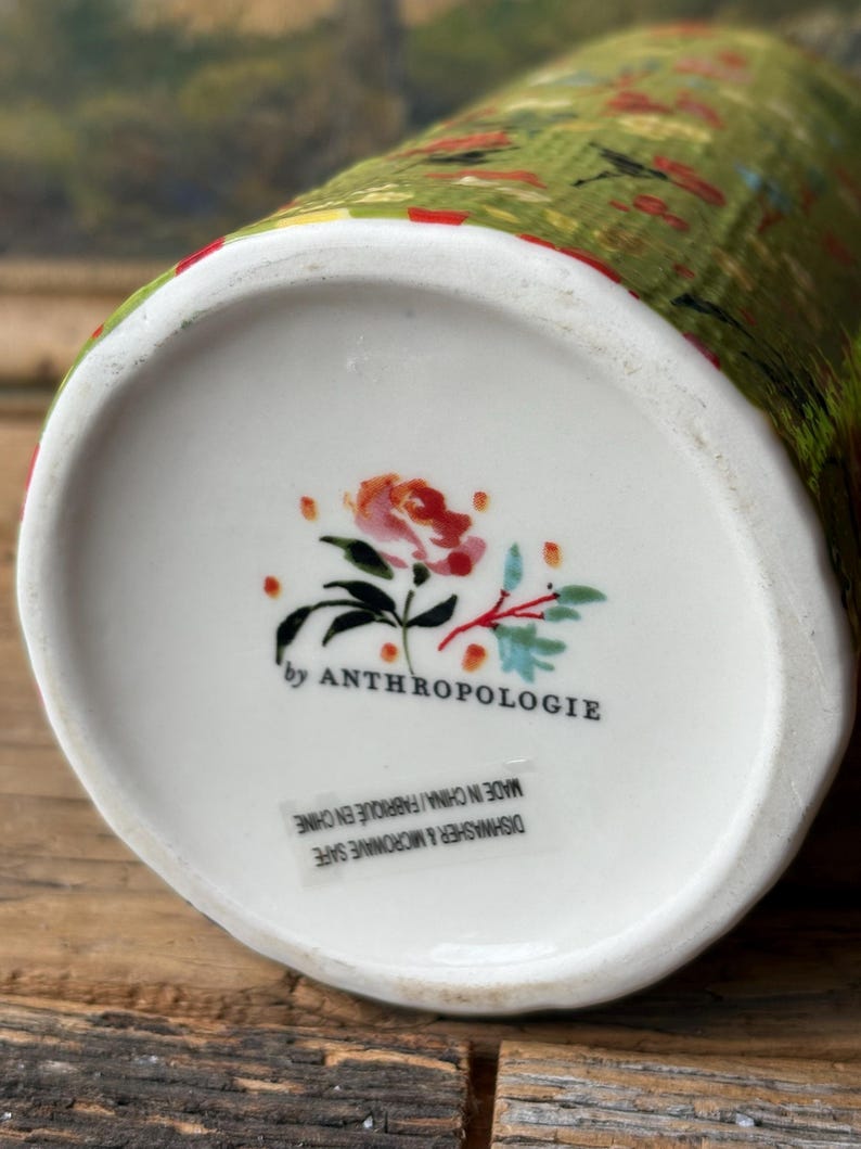 May include: A white ceramic vase with a green and red floral pattern. The vase is marked "by Anthropologie" and "Dishwasher & Microwave Safe. Made in China/Fabriqu&eacute; en Chine."