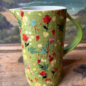 May include: A green ceramic pitcher with a floral pattern. The pitcher has a white rim and a green handle.