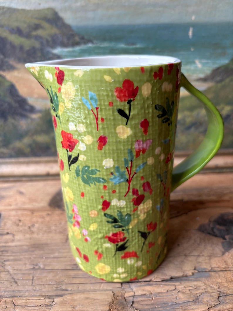 May include: A green ceramic pitcher with a floral pattern. The pitcher has a white rim and a green handle.
