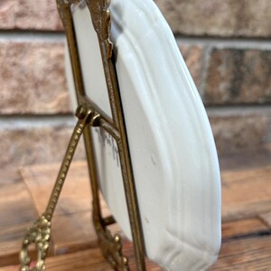 May include: A small, off-white ceramic plate displayed in a decorative, ornate gold-colored metal stand. The plate has a slightly curved shape and appears to have a subtle texture. The stand has a detailed, vintage design, with a hinged support.