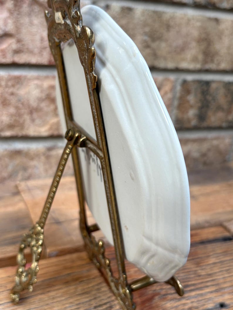 May include: A small, off-white ceramic plate displayed in a decorative, ornate gold-colored metal stand. The plate has a slightly curved shape and appears to have a subtle texture. The stand has a detailed, vintage design, with a hinged support.
