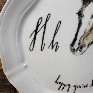 May include: White ceramic plate with a scalloped edge. The plate features a black script design of the letters "Hh" and the phrase "happy you're here". A detailed illustration of a horse's head is also present.