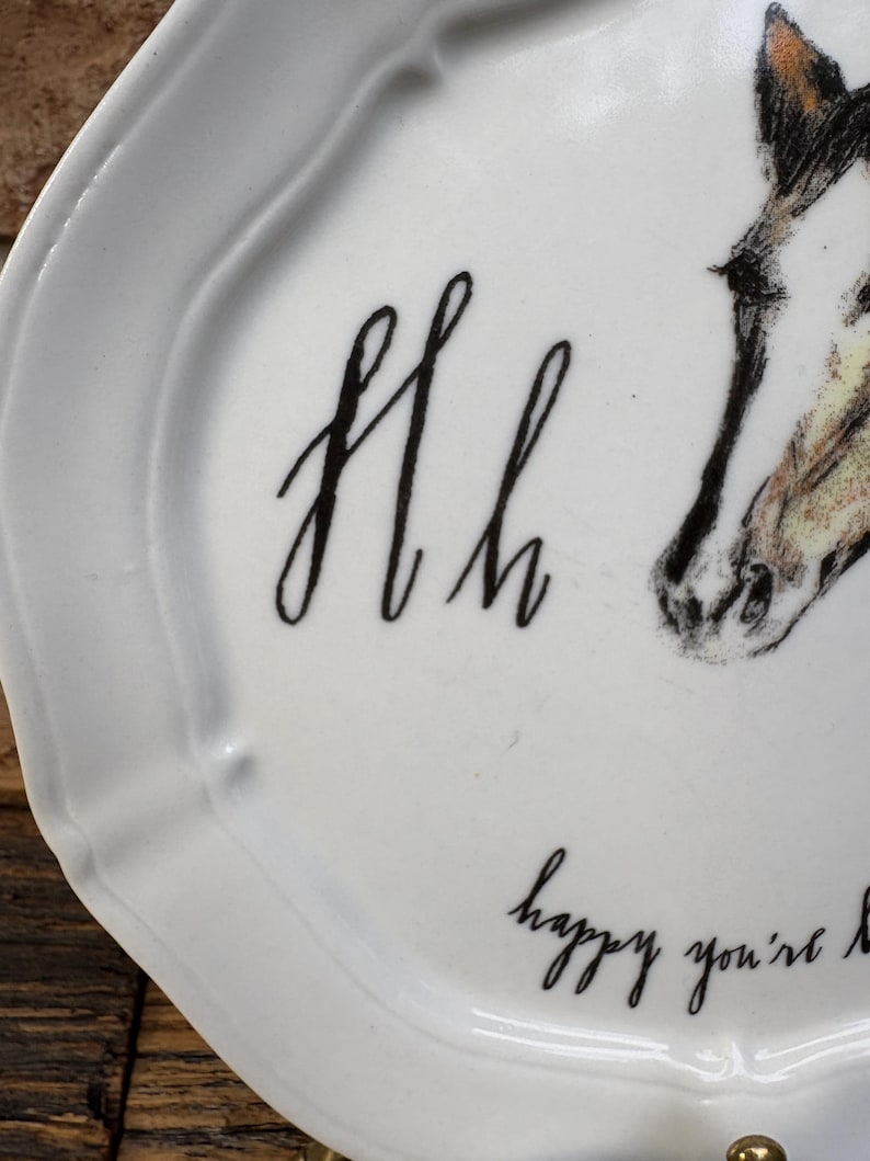 May include: White ceramic plate with a scalloped edge. The plate features a black script design of the letters "Hh" and the phrase "happy you're here". A detailed illustration of a horse's head is also present.