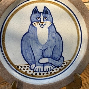 Puede incluir: Plato de cerámica con una ilustración de un gato azul. El gato está pintado en azul con detalles blancos, sentado sobre una línea punteada. El plato tiene un borde marrón y un borde dorado, con un fondo blanco moteado.