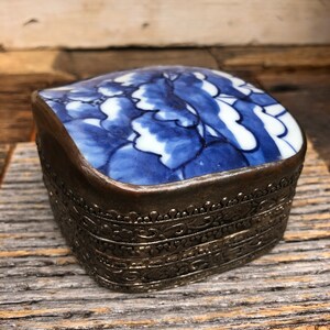 Puede incluir: Una pequeña caja cuadrada de plata con una tapa de porcelana azul y blanca. La tapa presenta un diseño floral con flores azules y hojas blancas.